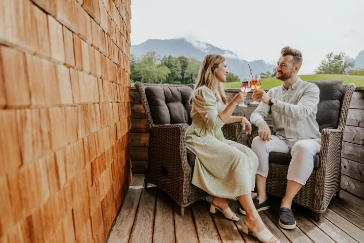 Ein Paar st&ouml;&szlig;t mit Drinks in einem gem&uuml;tlichen Au&szlig;enbereich des Hotel Klosterhof an, umgeben von Alpenlandschaft in Bayern.