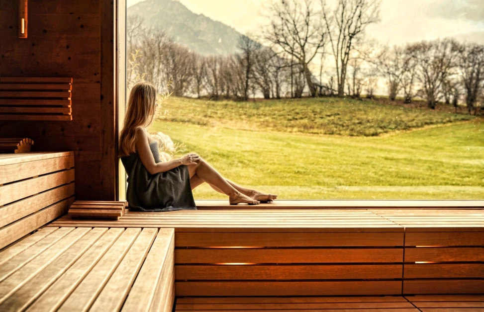 A woman in a towel relaxes on a wooden bench in a sauna, gazing at a scenic view of a grassy field and mountains through a large window.