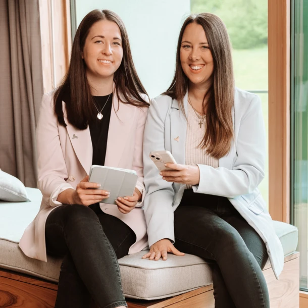 Two women sitting indoors, smiling, holding smartphones, with a window view of greenery.