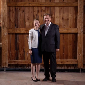 Klosterhof-Gastgeber, Henrike und Andreas Färber, posieren vor einer rustikalen Holzwand im Hotel Klosterhof, dem 4*S Wellnesshotel in Bayerisch Gmain, Bayern.