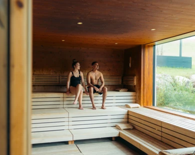 Ein Paar sitzt entspannt in einer modernen Sauna mit Holzverkleidung und großen Fenstern, die einen Blick ins Grüne bieten.