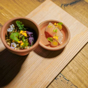 Ein kunstvoll angerichtetes Gericht mit essbaren Blumen und Kräutern in einem Hotel Klosterhof-Teller, umgeben von natürlichem Holz.