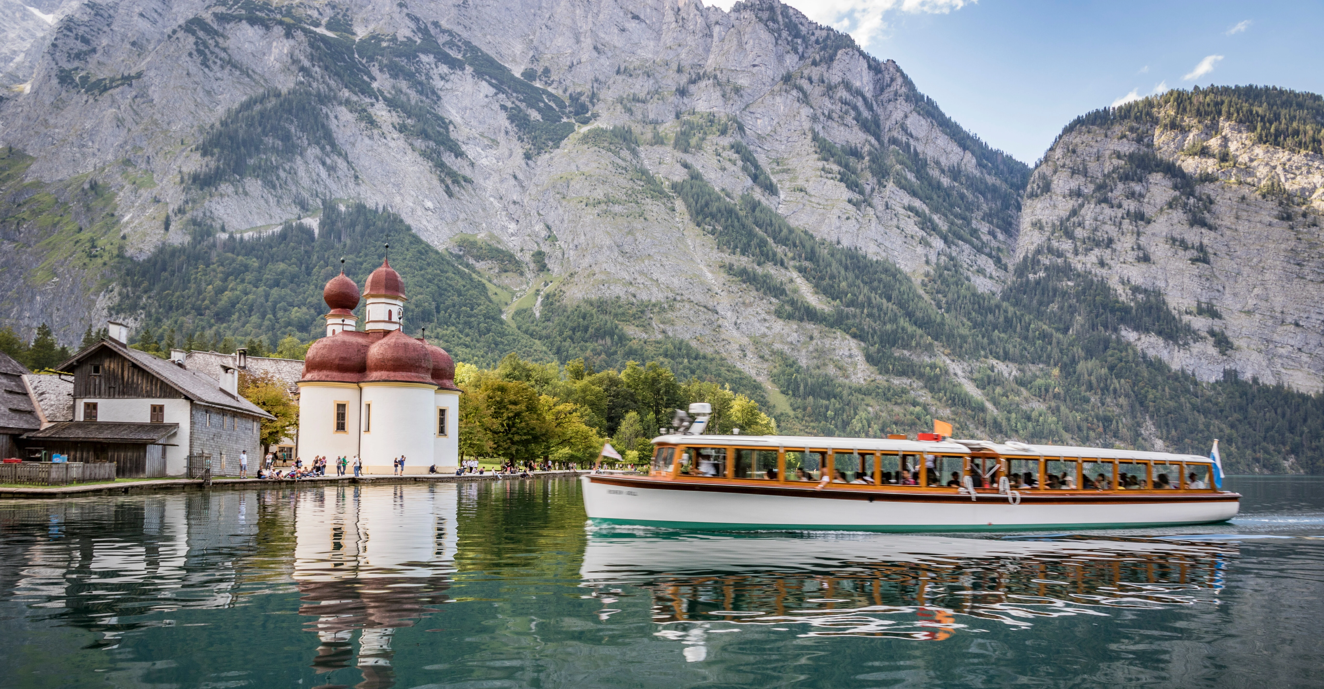 A boat glides across a serene lake with a picturesque church and mountains in the background.
