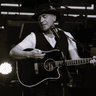 Musician playing acoustic guitar on stage, wearing a hat and vest, with stage lights in the background.