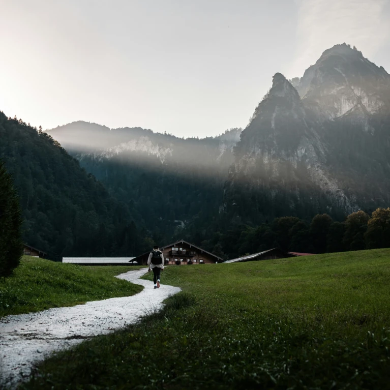 Eine Person wandert auf einem Pfad durch die grüne Wiese, umgeben von Bergen und dem Hotel Klosterhof im Hintergrund.