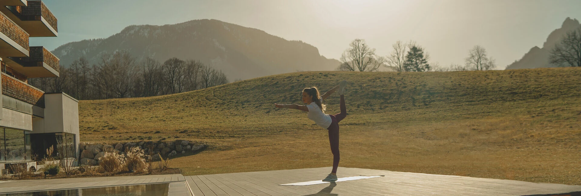 Eine Frau praktiziert Yoga auf einer Holzplattform mit Blick auf die Berge, neben einem Pool und einem Feuerplatz.