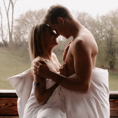 A couple smiling at each other, wrapped in a duvet, with a scenic view of trees in the background.
