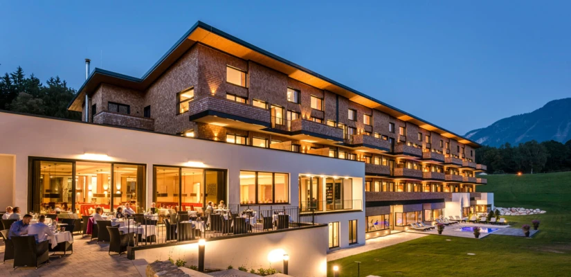 Modern hotel with outdoor dining area at dusk, surrounded by lush greenery and mountains.