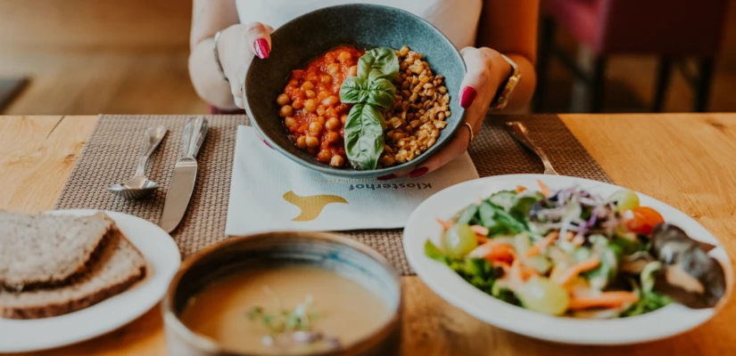 A person holding a bowl of chickpeas and grains with basil, alongside a salad, soup, and bread on a wooden table.