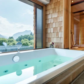 Blick auf eine Badewanne mit grünem Wasser und Aussicht auf Berge durch große Fenster in einem modernen Wellnessbereich.