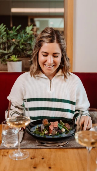 A woman enjoying a gourmet meal at Wellnesshotel Klosterhof in Bayern, with wine glasses and potted plants in the background.