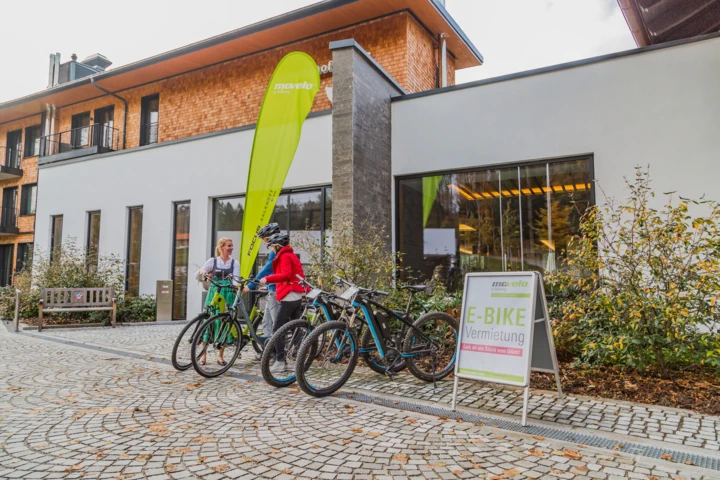 Zwei Personen stehen vor dem Hotel Klosterhof und besprechen E-Bike-Verleih. Im Hintergrund ist das moderne Gebäude und ein Werbeschild sichtbar.