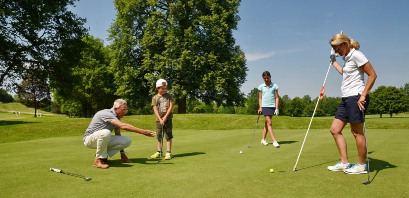 Ein Golftrainer zeigt einem Kind die richtige Technik, während zwei Mädchen aufmerksam zuschauen. Idylle im Hotel Klosterhof, Bayern.