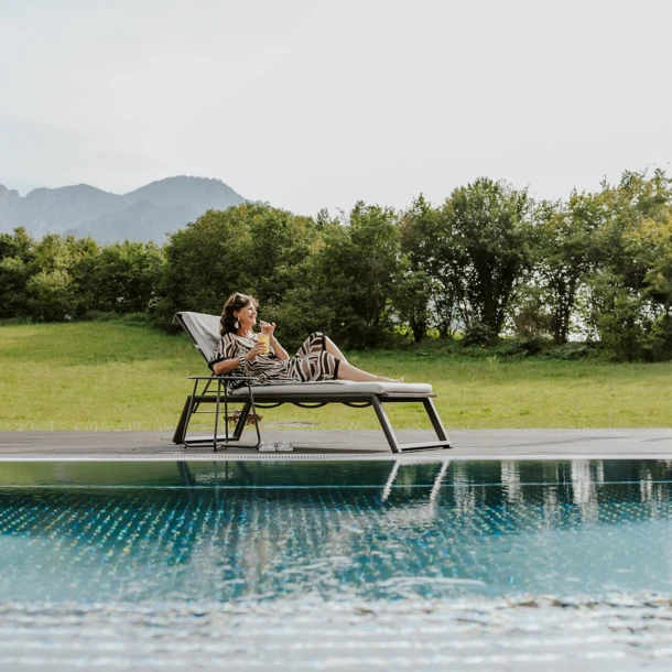 Eine Frau entspannt auf einer Liege am Pool, umgeben von grünen Wiesen und Bergen im Hintergrund.