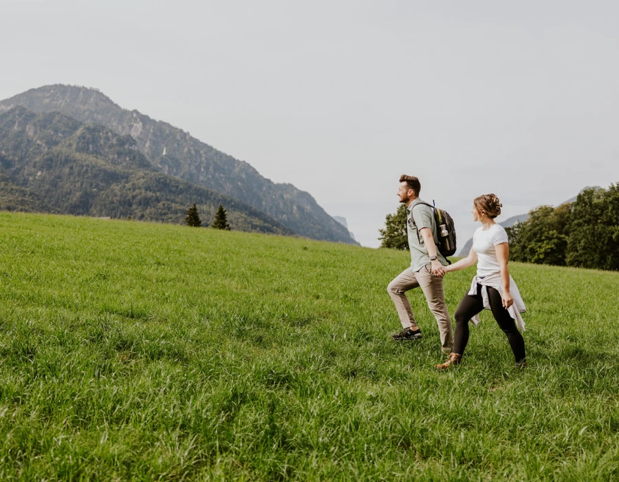Ein Paar wandert Hand in Hand &uuml;ber eine gr&uuml;ne Wiese mit Bergen im Hintergrund.
