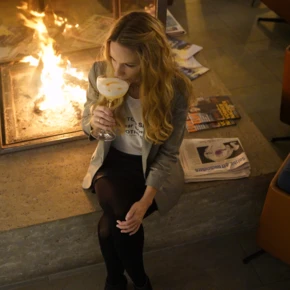 A woman enjoys a drink by the fireplace at Hotel Klosterhof, with magazines and newspapers nearby, creating a cozy atmosphere.