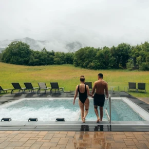 A couple entering an outdoor pool at Wellnesshotel Klosterhof, surrounded by lush greenery and misty mountains.