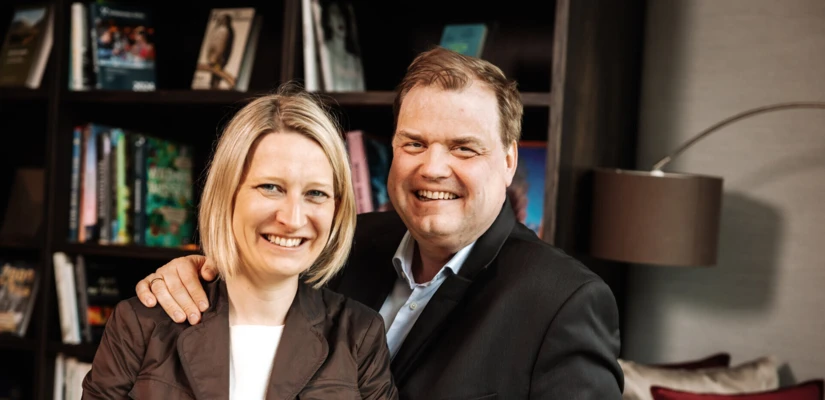 Ein l&auml;chelndes Paar posiert entspannt in einem modernen Innenraum des Hotel Klosterhof, umgeben von B&uuml;cherregalen.