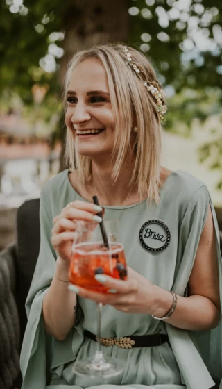 Eine lächelnde Frau in einem grünen Kleid hält ein Getränk mit Beeren. Sie trägt ein "Bride"-Abzeichen und sitzt entspannt im Freien.