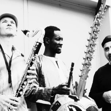 Drei Musiker mit Instrumenten, darunter Klarinette und Kora, vor einer Wand.