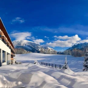 Snowy landscape with a modern hotel, mountains, and trees under a bright blue sky. Sunlight reflects on the snow, creating a serene winter scene.