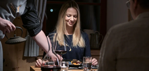 Eine Frau genießt ein feines Dinner, während ein Kellner ihr Essen serviert, umgeben von gemütlicher Atmosphäre und Kerzenlicht.