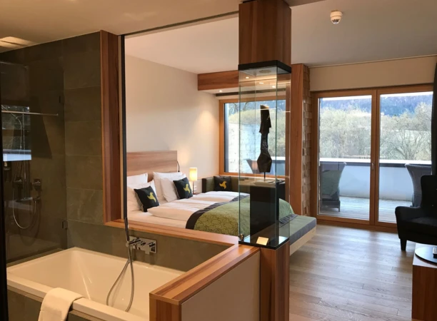 Modern hotel room with a cozy bed, elegant bath area, and a view of the greenery through large windows.