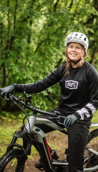Frau mit Helm lächelt auf ihrem E-Bike in einer grünen Umgebung, bereit für ein Abenteuer in der Natur.