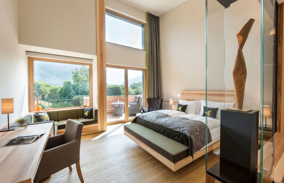 Modern hotel room with large windows offering mountain views, featuring a bed, desk, seating area, and balcony. Wellnesshotel Klosterhof in Bayern.