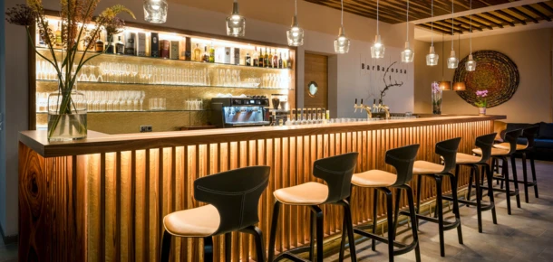 Modern bar with wooden decor, high stools, and pendant lights at Klosterhof. Shelves behind display glasses and bottles, creating a cozy atmosphere.