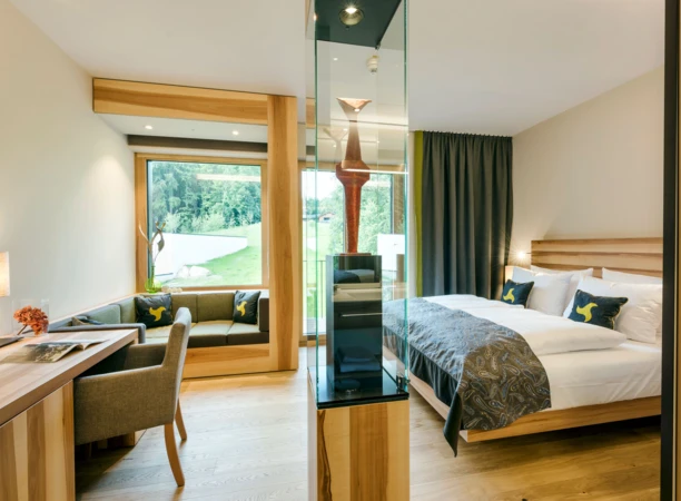 Modernes Hotelzimmer mit großem Bett, gemütlicher Sitzecke und Blick ins Grüne durch ein großes Fenster. Elegantes und einladendes Design.