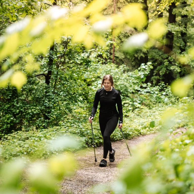 Eine Frau wandert mit Stöcken auf einem Waldweg, umgeben von üppigem Grün und Bäumen.