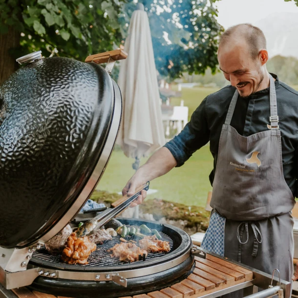 Ein Koch grillt Fleisch und Gemüse auf einem großen Grill im Freien, umgeben von einer schönen, grünen Landschaft.