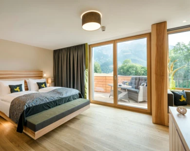 Modernes Hotelzimmer mit gro&szlig;em Bett, Holzboden und gro&szlig;er Fensterfront. Terrasse mit Sitzm&ouml;beln und Blick auf die Berge im Hintergrund.