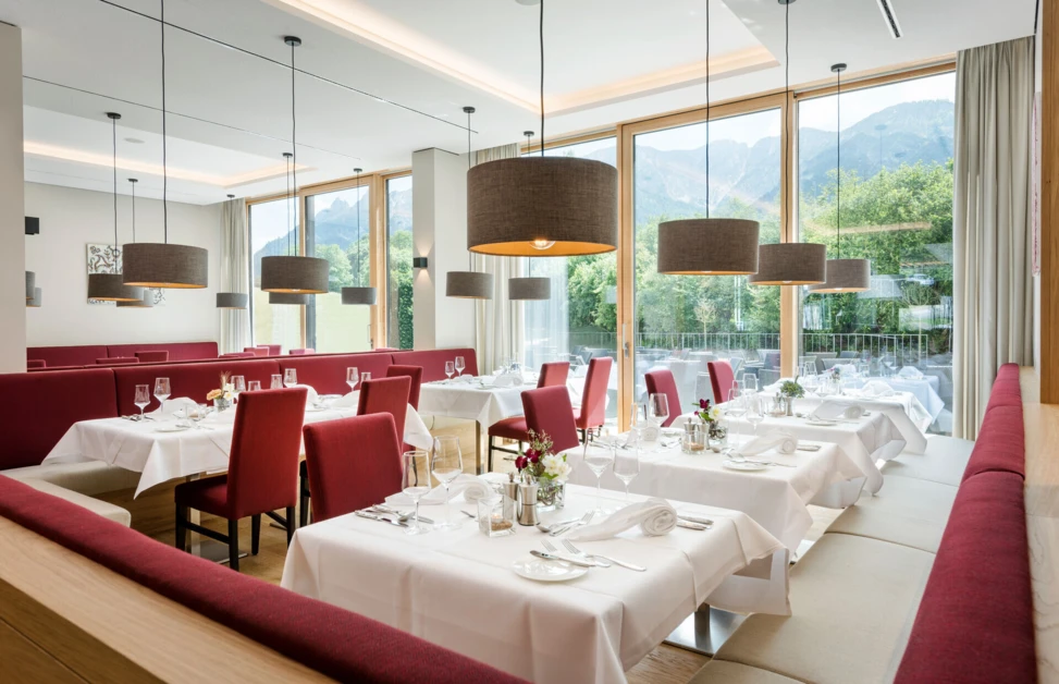 Elegant dining area with red chairs and white tablecloths at Hotel Klosterhof, featuring large windows with mountain views.