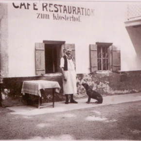 Ein Mann in Schürze steht vor dem Café Restauration zum Klosterhof, neben einem Hund und einem Tisch. Historische Szenerie in Bayern.