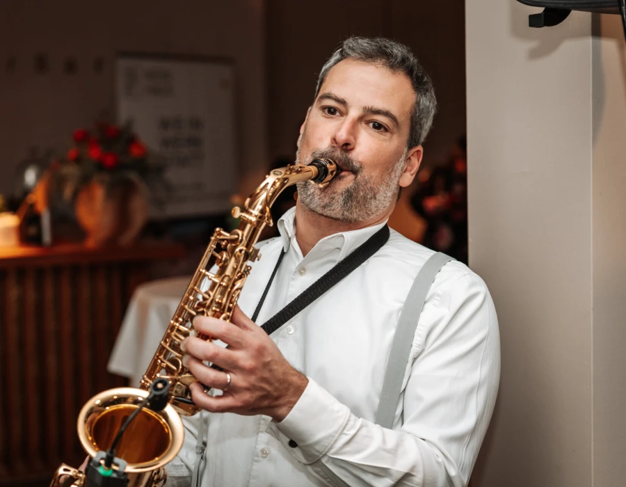 Ein Musiker spielt Saxophon in einem stilvollen Ambiente, w&auml;hrend G&auml;ste im Hintergrund entspannen.