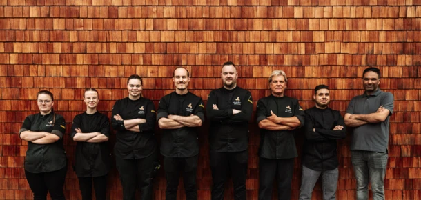Ein Team von Köchen steht lächelnd vor einer Holzfassade und zeigt stolz ihre Kochuniformen.