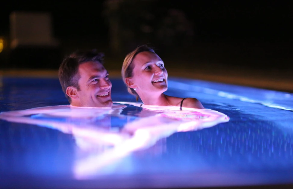A couple enjoys a night swim in a pool with glowing lights, creating a romantic and relaxing atmosphere.