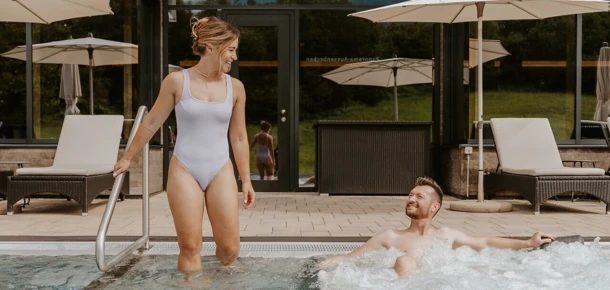 Eine Frau in einem weißen Badeanzug steht am Poolrand, während ein Mann im Wasser entspannt. Hintergrund zeigt das Hotel Klosterhof in Bayern.