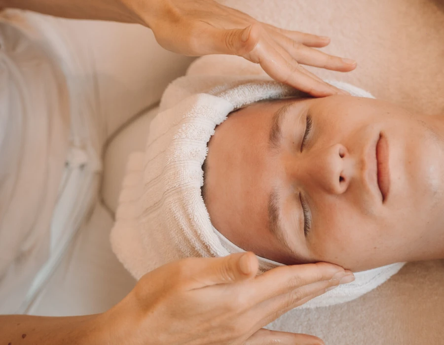 Ein junger Mann erhält eine entspannende Gesichtsbehandlung mit einem Handtuch um den Kopf in einer Wellnessumgebung.