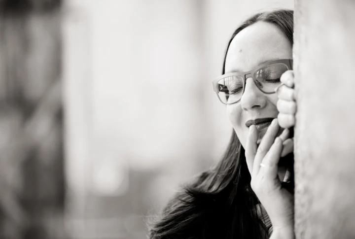 Eine Frau mit Brille lächelt sanft und lehnt sich an eine Wand, in einem entspannten Moment.