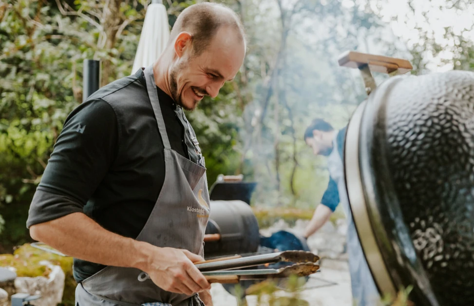 Ein Koch lächelt, während er mit Zangen am Grill in einem Garten arbeitet, umgeben von Bäumen und Pflanzen.