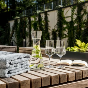 Towels, glasses, and a book on a wooden table outdoors, with greenery in the background.