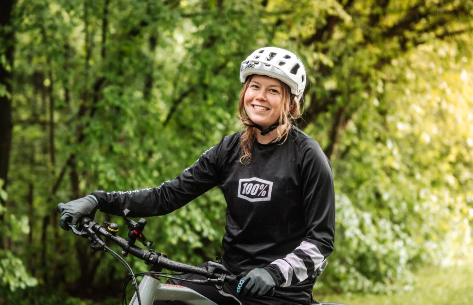 Eine lächelnde Frau in einem Radhelm sitzt auf ihrem Fahrrad in einer grünen Waldumgebung.