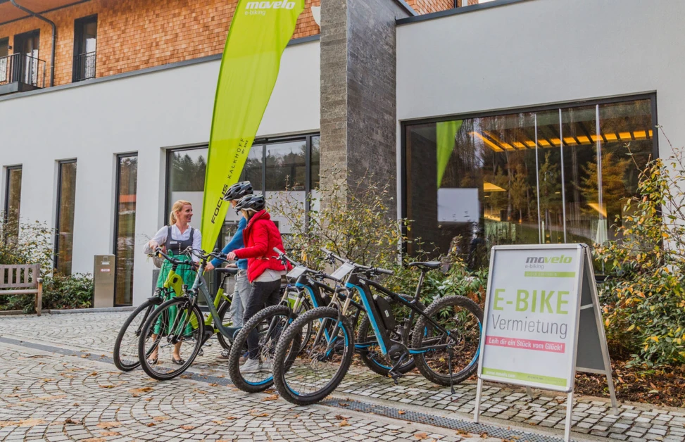 Zwei Personen stehen vor dem Hotel Klosterhof und besprechen E-Bike-Verleih. Im Hintergrund ist das moderne Gebäude und ein Werbeschild sichtbar.