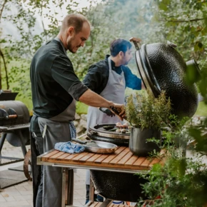 Zwei Köche grillen im Freien an einem großen Grill, umgeben von grüner Vegetation.