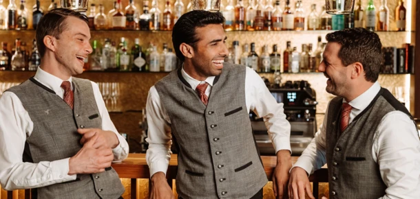 Drei lächelnde Barkeeper in eleganter Kleidung stehen an einer Bar, umgeben von Spirituosenflaschen im Hotel Klosterhof.