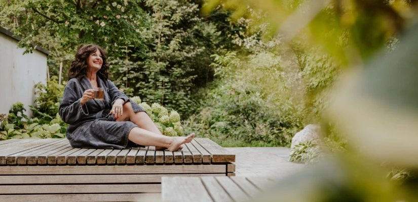Frau in einem Bademantel sitzt entspannt auf einer Holzliege und genießt eine Tasse Tee in einer grünen Gartenlandschaft.