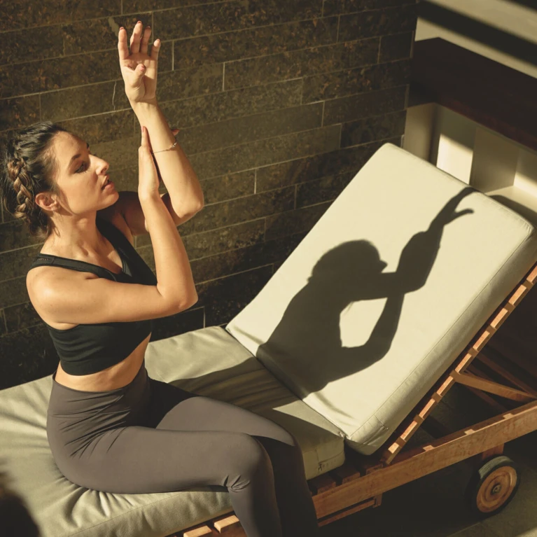 A woman in yoga attire sits on a lounge chair, practicing shadow hand gestures against a sunlit wall.